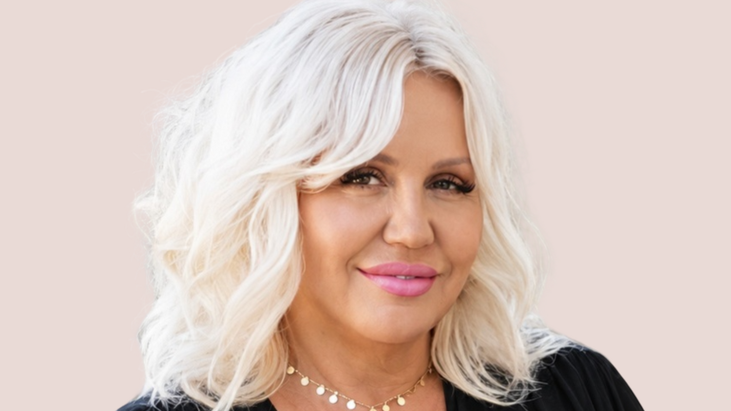A esthetician with shoulder-length platinum blonde hair wearing a black draped V-neck blouse and a delicate gold necklace, set against a solid pale peach background.